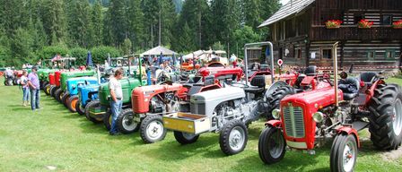 Mehrere Traktoren sind in einem Feld neben einem Holzschuppen geparkt. Die Traktoren sind in verschiedenen Farben und einige haben Anhänger. Eine Person steht neben einem roten Traktor.