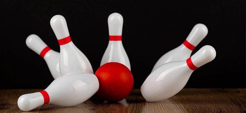 Ein roter Bowlingball wirft weiße Bowlingkegel auf einem Holztisch um, vor einem schwarzen Hintergrund.