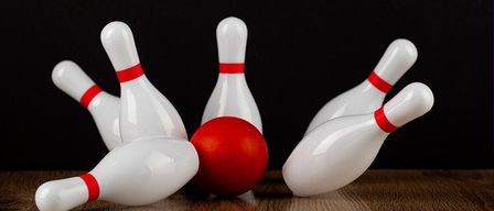 Ein roter Bowlingball wirft weiße Bowlingkegel auf einem Holztisch um, vor einem schwarzen Hintergrund.