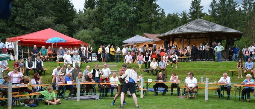 Zwei Männer ringen auf einem Rasenfeld. Zuschauer sehen von den Tribünen und Tischen zu. Zelte und Regenschirme befinden sich an der Seite. Bäume und Häuser sind in der Ferne.