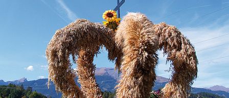 Ein Bild eines großen Herzens aus getrockneten Weizenhalmen mit zwei Sonnenblumen darauf. Dahinter steht ein Kreuz gegen einen blauen Himmel, mit Bergen in der Ferne.