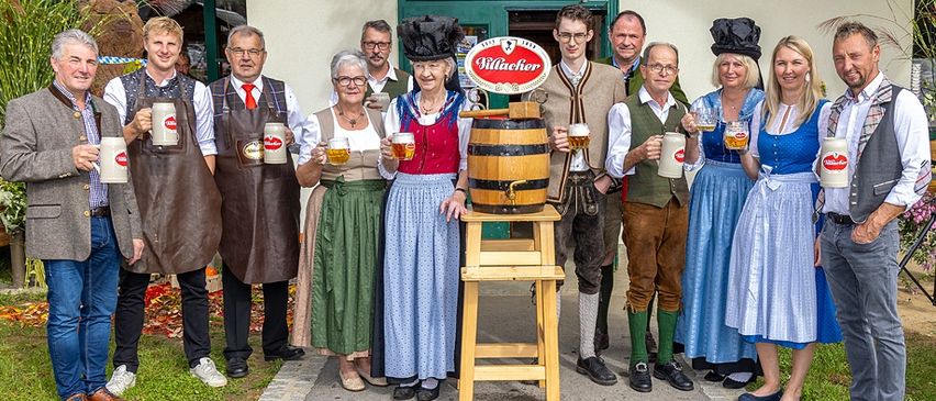 Eine Gruppe von Menschen in traditioneller deutscher Kleidung steht zusammen und hält Bierkrüge. Ein Fass steht auf einem Hocker in der Mitte der Gruppe.