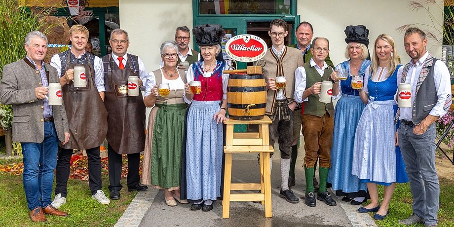 Eine Gruppe von Menschen in traditioneller deutscher Kleidung steht zusammen und hält Bierkrüge. Ein Fass steht auf einem Hocker in der Mitte der Gruppe.