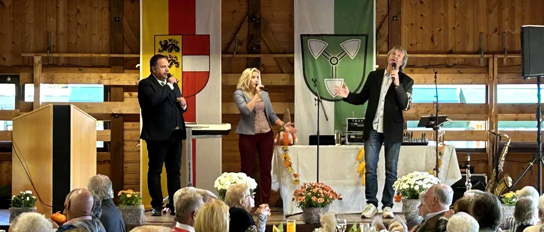 Drei Personen stehen auf einer Bühne vor einem Publikum. Zwei von ihnen halten Mikrofone und eine hält eine Kürbis. Hinter ihnen sind Banner mit Wappen. Das Publikum sitzt an einem Tisch mit Gläsern und Tellern.