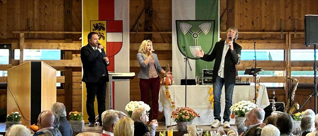 Drei Personen stehen auf einer Bühne vor einem Publikum. Zwei von ihnen halten Mikrofone und eine hält eine Kürbis. Hinter ihnen sind Banner mit Wappen. Das Publikum sitzt an einem Tisch mit Gläsern und Tellern.