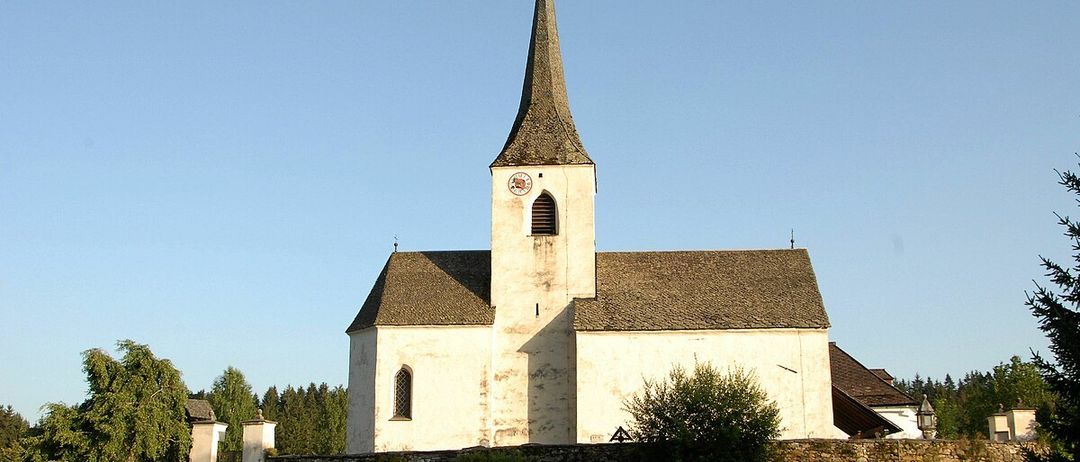 Eine Kirche mit einem spitzen Turm und einer Uhr steht auf einem Hügel unter einem klaren Himmel.