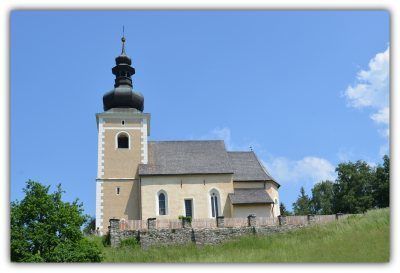 Eine traditionelle Kirche mit einem Kirchturm, umgeben von einer Steinmauer, auf einer grasbewachsenen Anhöhe unter einem klaren blauen Himmel.