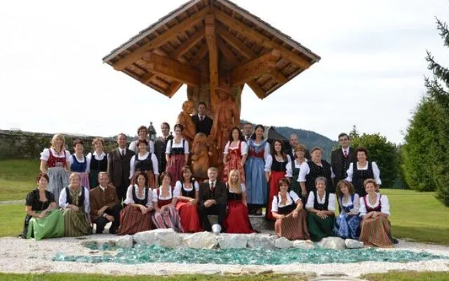 Eine Gruppe von Menschen in traditioneller Kleidung posiert für ein Foto vor einer Holzstruktur mit einer Statue, umgeben von Steinen und Gras in einem Außenbereich.