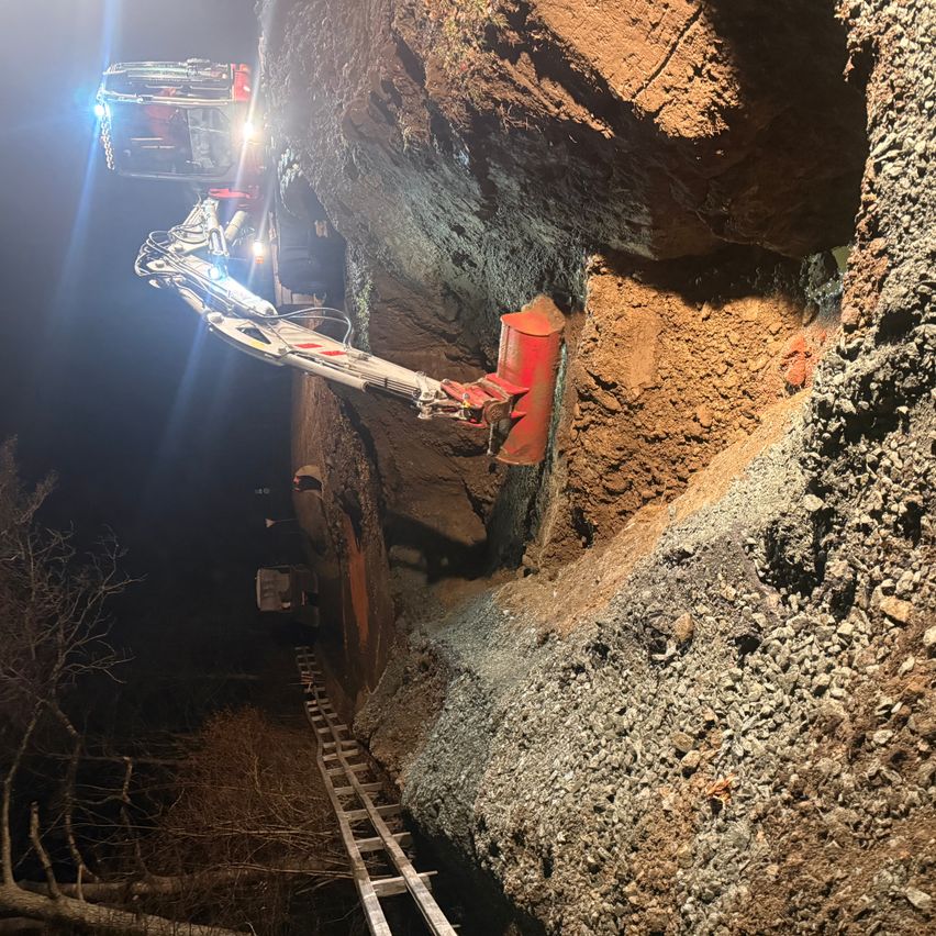 Ein Baufahrzeug gräbt nachts in einen Fels. Das Fahrzeug hat einen roten Anbau und wird von einem blauen Licht beleuchtet. Eine Leiter ist sichtbar, die in den Fels hinunterführt.