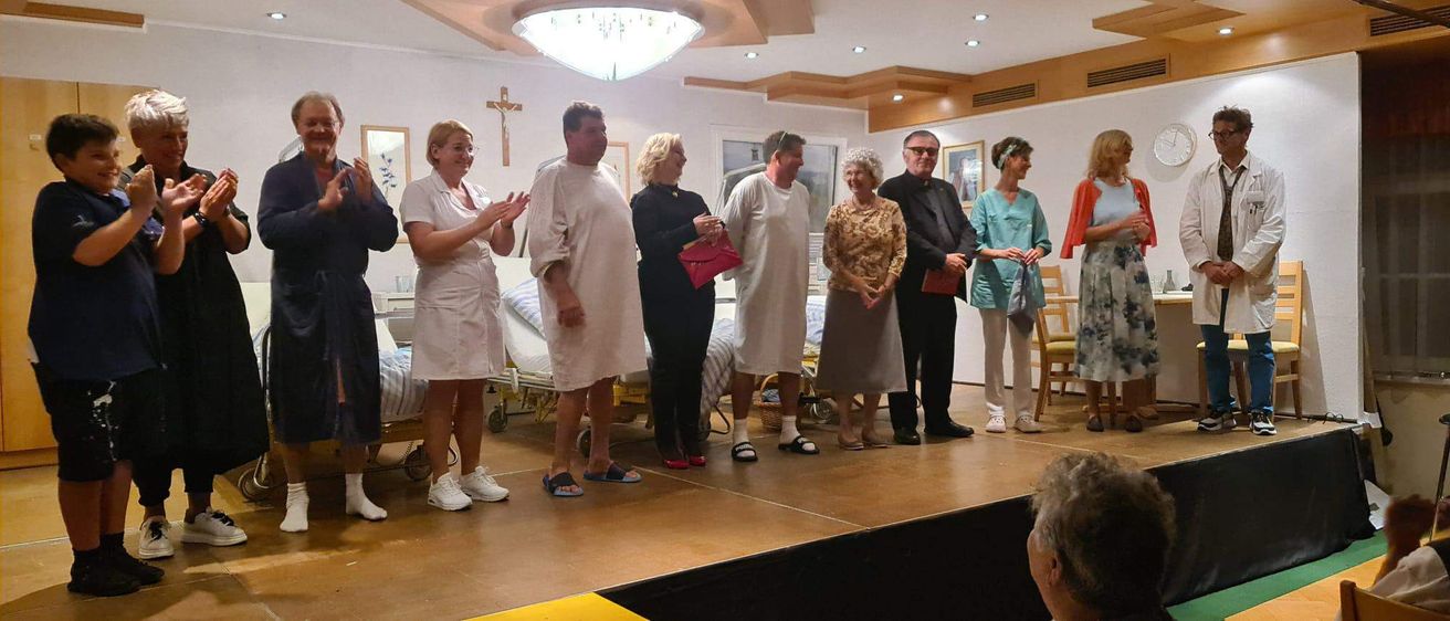 Eine Gruppe von Menschen in Krankenhauskittel stehen auf einer Bühne mit einem Priester und zwei Frauen in Scrubs, applaudieren. Dahinter stehen zwei Betten mit Patienten und ein Krankenhauszimmer mit gerahmten Bildern.