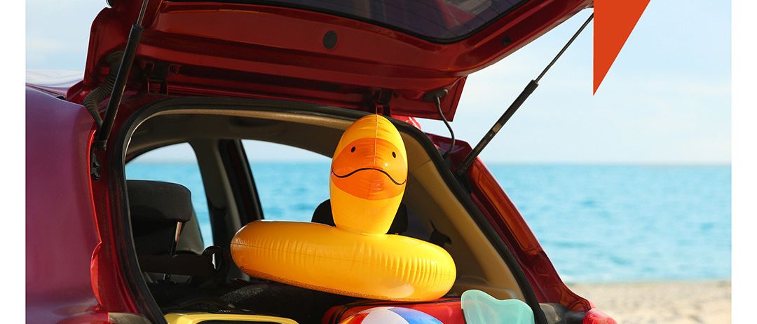 Ein rotes Auto mit offenem Kofferraum, gefüllt mit Strandutensilien wie Ball, aufblasbarem Schwimmring und Schnorcheln. Der Kofferraum befindet sich an einem Strand mit Meer im Hintergrund. Der Text lautet 'Pack die Badehose ein...'.
