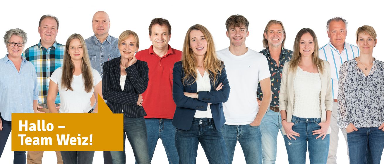 Fünf Menschen stehen in einer Reihe und lächeln. Eine Frau in der Mitte hat die Arme verschränkt, und ein Mann neben ihr trägt ein weißes T-Shirt. Dahinter tragen eine Frau und ein Mann Jeansjacken.