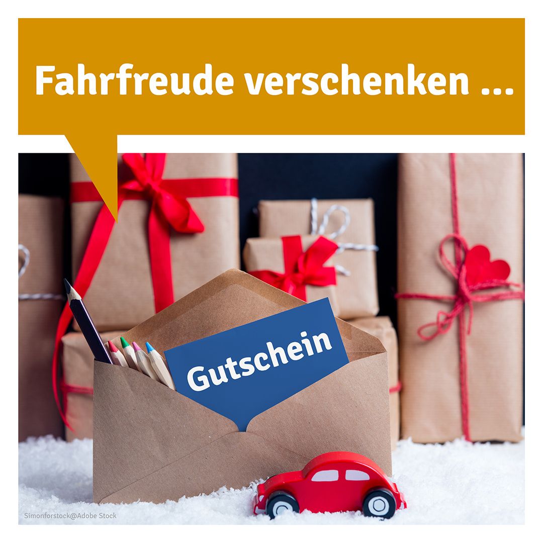 Ein rotes Auto steht neben einem offenen braunen Umschlag mit der Aufschrift 'Gutschein'. Dahinter befinden sich mehrere verpackte Geschenke mit roten Bändern und Herzen. Der Hintergrund lautet 'Fahrfreude verschenken...'
