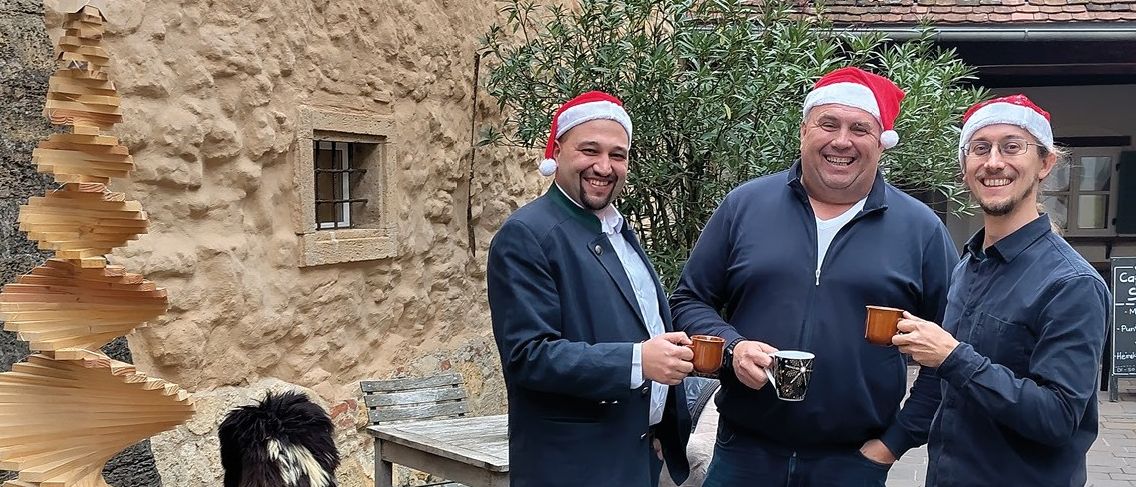 Drei Männer in festlichen Mützen stehen vor einem Gebäude und halten Tassen in der Hand. Neben ihnen steht ein Holz-Weihnachtsbaum. Die Veranstaltung trägt den Titel 'Weihnachteln in der Saxxess Passage'. Die Veranstaltung läuft vom 25. November bis 18. Dezember 2025. Sie ist jeden Dienstag, Mittwoch und Donnerstag von 16 bis 22 Uhr geöffnet.