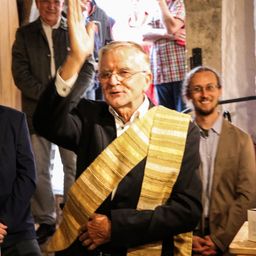 Ein älterer Mann mit einem goldenen Schal winkt der Menge zu, während er neben einem anderen Mann mit Brille steht. Dahinter steht eine Gruppe von Menschen und beobachtet.