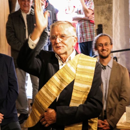 Ein älterer Mann mit einem goldenen Schal winkt der Menge zu, während er neben einem anderen Mann mit Brille steht. Dahinter steht eine Gruppe von Menschen und beobachtet.