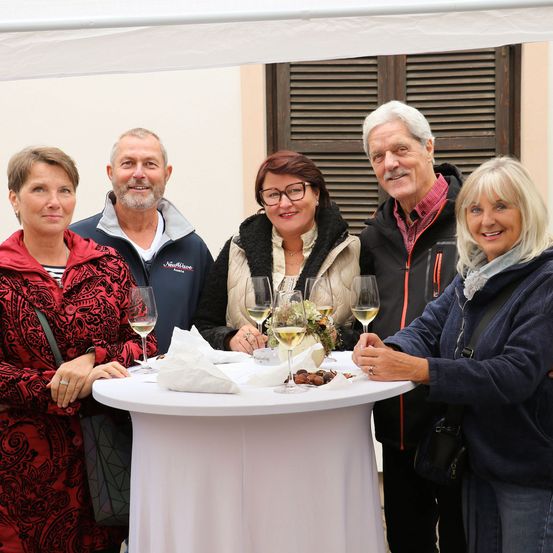 Eine Gruppe von sechs Personen, darunter vier Frauen und zwei Männer, stehen lächelnd zusammen unter einem Zelt. Sie haben sich um einen Tisch mit Weingläsern und einem Blumenstrauß versammelt.