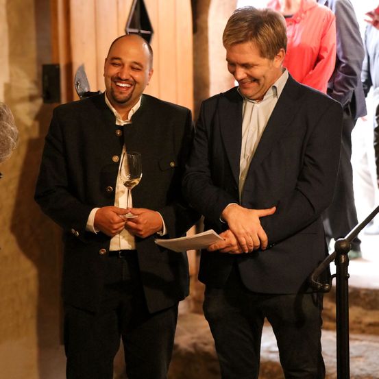 Zwei Männer in formeller Kleidung, die lächeln und ein Weinglas halten, stehen in der Nähe einer Treppe. Ein Papier befindet sich in der rechten Hand des Mannes auf der rechten Seite. Sie sind von Menschen umgeben.
