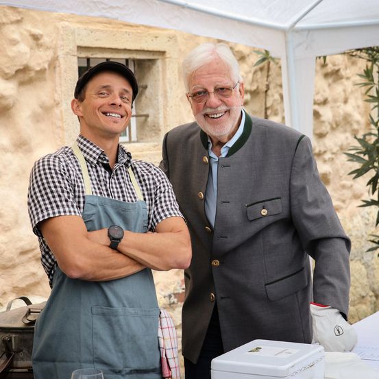 Zwei Männer stehen draußen, einer in einem grauen Anzug und der andere in einem karierten Hemd mit einer Schürze. Sie lächeln beide und posieren für ein Foto vor einer Steinmauer.