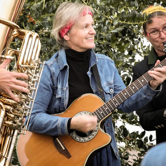 Eine Frau mit einem roten Stirnband spielt lächelnd eine Gitarre. Daneben steht eine Person, die eine Trompete spielt, und ein Mann mit Brille spielt Klarinette. Sie stehen vor einer Pflanze mit grünen Blättern.