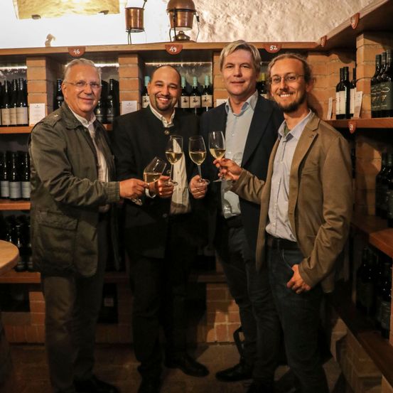 Vier Männer stehen in einem Weinkeller, lächeln und halten Weingläser in den Händen. Hinter ihnen sind mit Weinflaschen gefüllte Regale zu sehen, wobei einige Etiketten Zahlen und Text anzeigen.