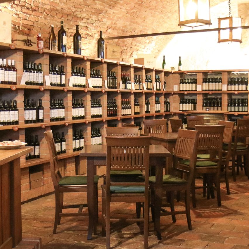 Innenansicht eines Weinkellers mit Holzregalen voller Weinflaschen. Es gibt Holztische und Stühle in einer Reihe, mit einem Fass im Hintergrund. Eine Lampe hängt von der Decke.