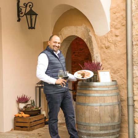 Ein Mann hält einen Teller mit Essen und zwei Weingläser in der Hand und lächelt vor einem Fass und einer Steinmauer.