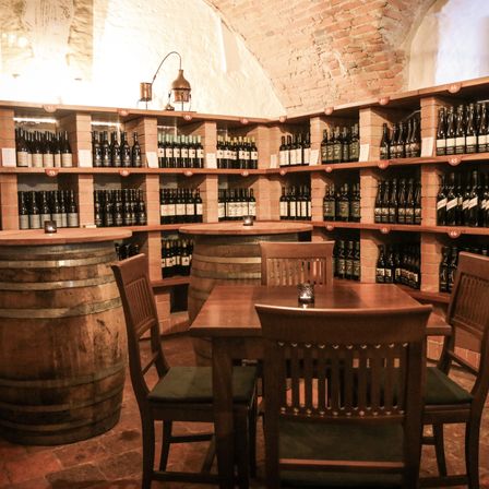 Ein Weinkeller mit Ziegelwänden, Holzfässern und Regalen voller nummerierter Weinflaschen. Vier Holzstühle umgeben einen Tisch mit einer Kerze darauf.