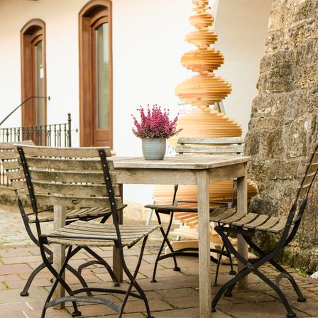 Ein Esstisch im Freien mit einer Blumenvase darauf, umgeben von Holzstühlen. Dahinter steht eine hölzerne Spiralskulptur und ein Gebäude mit Rundbogenfenstern.