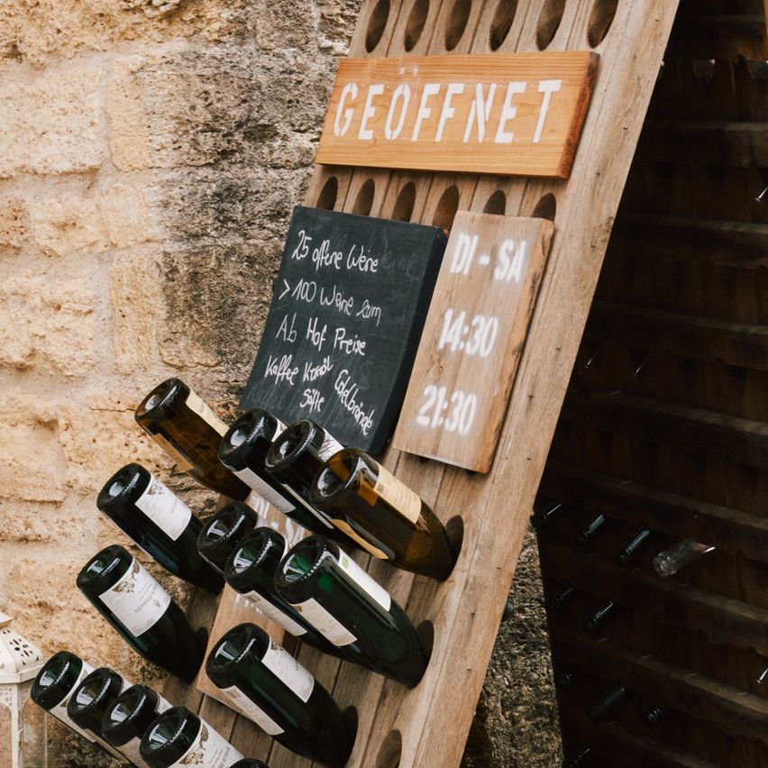 A wooden rack with wine bottles and a sign reading 'Geoffnet'. Below the sign is a chalkboard with wine offerings and times.