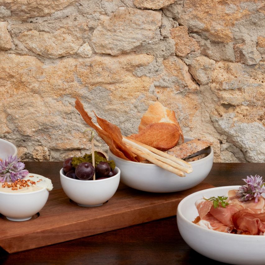 A wooden tray with various bowls of food on it, including a bowl of olives, a bowl of cheese, and a bowl of breadsticks.