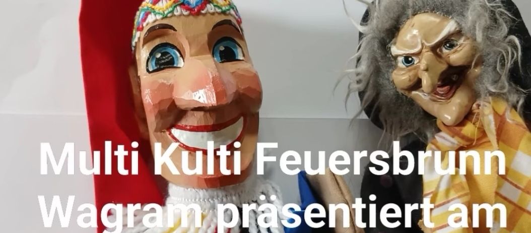 Zwei Puppen stehen vor einem weißen Brett mit buntem Text. Es steht: 'Multi Kultur Feuersbrunn Wagram präsentiert am 20. Juni 26 um 15 Uhr für Groß und Klein die Kremser Puppen Truhe.'