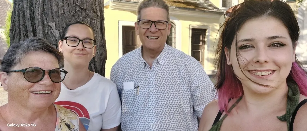 Vier Menschen lächeln vor einem Baum. Die Frau auf der rechten Seite hat pinkfarbene Haare und trägt ein grünes Oberteil. Der Mann in der Mitte trägt eine Brille. Die Frau auf der linken Seite hat graue Haare und trägt eine Sonnenbrille.