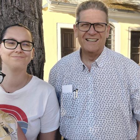 Drei Menschen stehen draußen. Ein Mann mit Brille und gemustertem Hemd lächelt in die Kamera. Daneben steht eine Frau in einem weißen T-Shirt und Brille. Hinter ihnen ist ein Baumstamm und ein gelbes Gebäude mit Fenstern zu sehen.