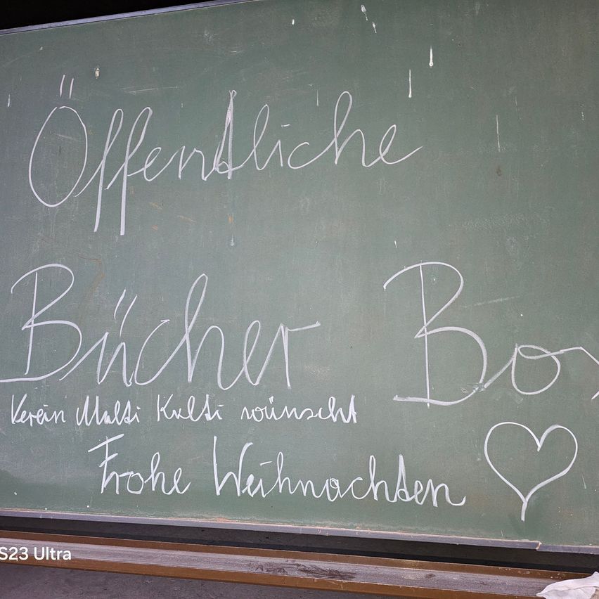Eine Tafel mit den Worten 'Offentlich Bucher Box' in weißer Kreide geschrieben. Darunter befindet sich ein Herz und 'Frohe Weihnachten'. Das Bild hat einen Wasserzeichen 'S23 Ultra'.