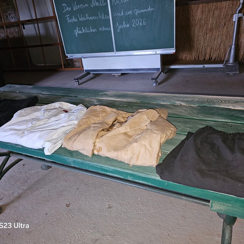 Eine grüne Bank mit drei Kleidungsstücken: einem weißen Hemd, einer beigen Jacke und einem schwarzen Rock. Hinter der Bank eine Tafel mit deutschem Text und einem Wasserzeichen 'S23 Ultra'.
