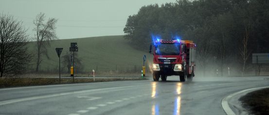 Ein roter Feuerwehrwagen mit blauen Lichtern fährt bei Regen auf einer nassen Straße. Bäume und Hügel befinden sich an den Seiten der Straße.