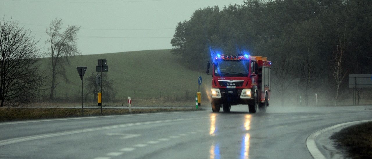 Ein roter Feuerwehrwagen mit blauen Lichtern fährt bei Regen auf einer nassen Straße. Bäume und Hügel befinden sich an den Seiten der Straße.