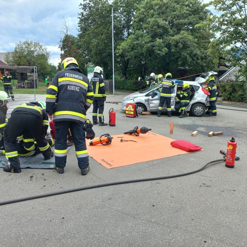 Feuerwehrleute in Schutzkleidung arbeiten an einem beschädigten Auto, mit Werkzeug und Ausrüstung auf dem Boden verteilt.