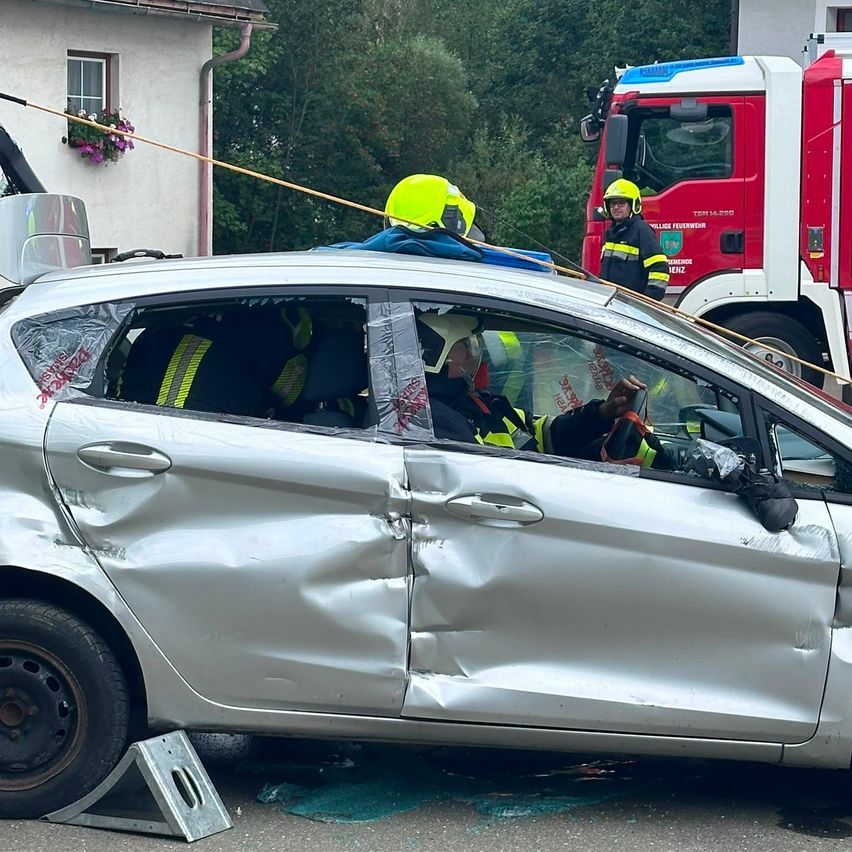 Ein silbernes Auto mit erheblichen Schäden wird von Notfallhelfern in gelben Helmen versorgt. Ein Feuerwehrwagen ist hinter dem Auto geparkt.