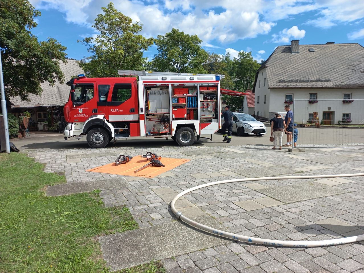 Ein roter Feuerwehrwagen ist auf einer gepflasterten Straße geparkt, mit offenen Türen und sichtbarer Ausrüstung. In der Nähe stehen Menschen, und ein weißes Auto ist dahinter geparkt. Die Szene spielt sich in einem Dorf ab, mit Häusern und Bäumen im Hintergrund.