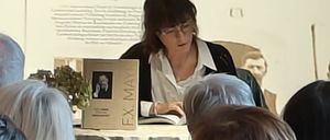 Eine Frau liest aus einem Buch an einem Tisch, umgeben von einer Gruppe älterer Erwachsener, die zuhören. Ein Bild von einem Haus und Menschen hängt an der Wand hinter ihr.