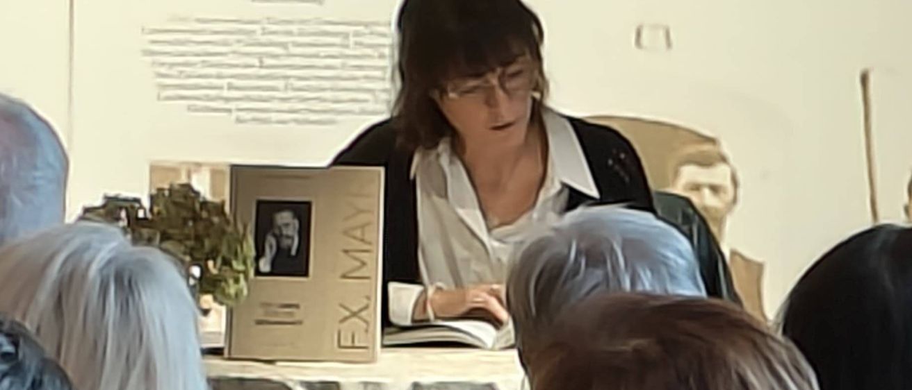 Eine Frau liest aus einem Buch an einem Tisch, umgeben von einer Gruppe älterer Erwachsener, die zuhören. Ein Bild von einem Haus und Menschen hängt an der Wand hinter ihr.