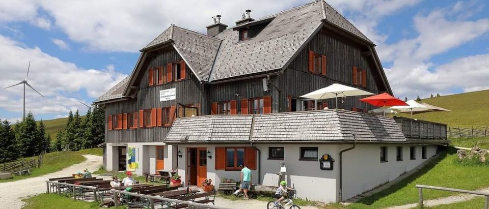 Ein Chalet-Stilgebäude mit roten Fensterläden und einem Kind, das vor dem Gebäude Fahrrad fährt. Eine Gruppe von Menschen sitzt auf Bänken in der Nähe des Gebäudes.