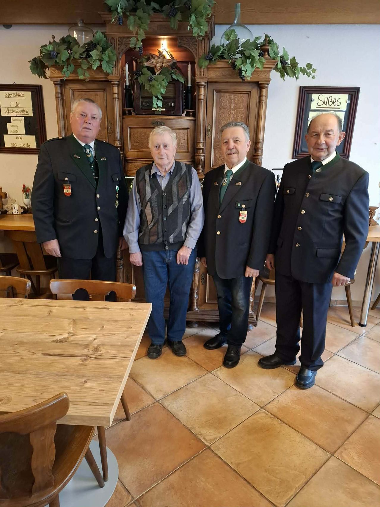 Vier Männer stehen zusammen in einem Raum, wahrscheinlich bei einer gesellschaftlichen Veranstaltung. Ein Mann trägt einen dunklen Anzug mit einem grünen Schal.