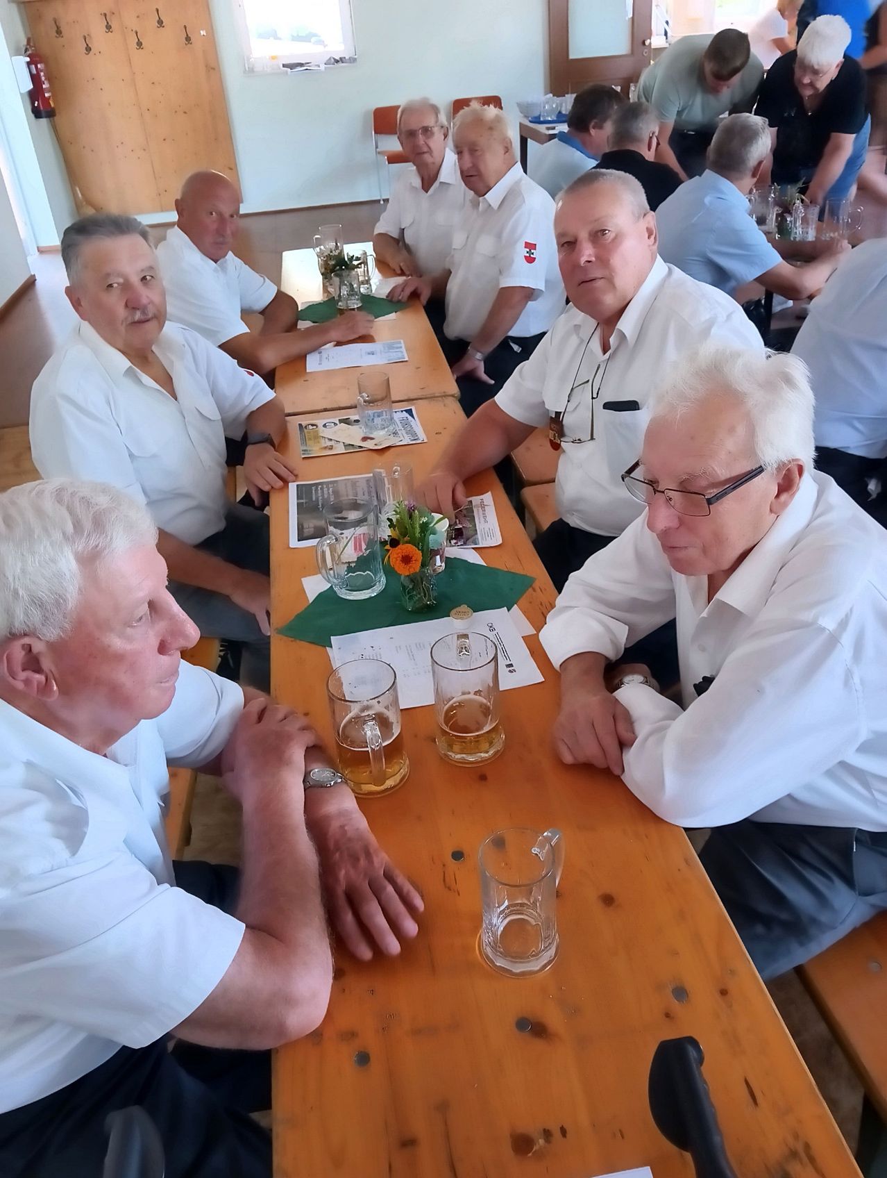 A group of older men in white shirts are sitting at a long table, possibly in a restaurant, with glasses of beer and a flower centerpiece.