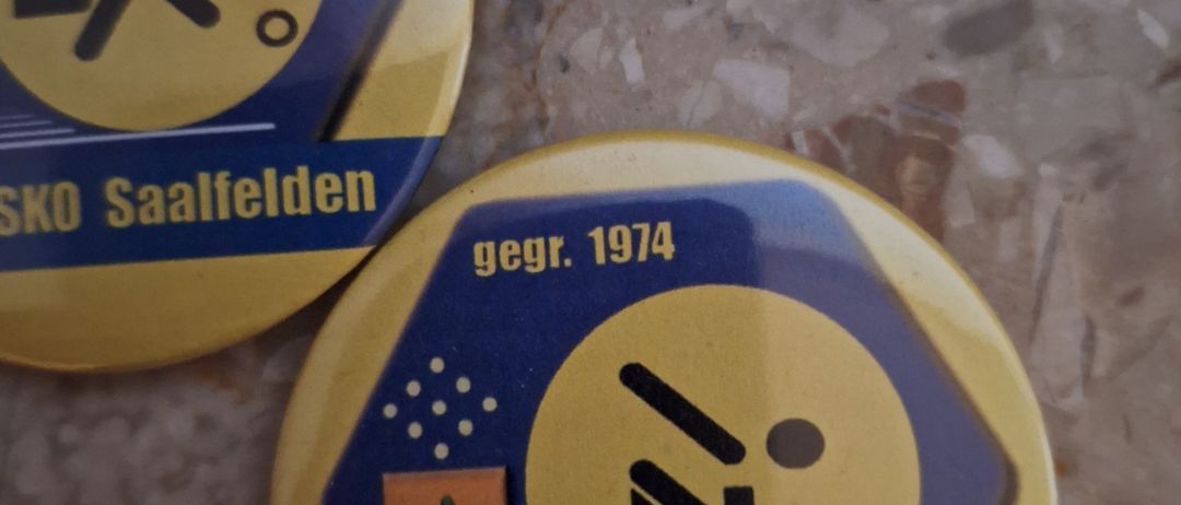 Zwei gelbe und blaue sechseckige Pins mit einer laufenden Figur und den Worten '1. KC ASKO Saalfelden' darauf. Ein Pin hat ein kleines Logo links und das Jahr 1974 rechts.
