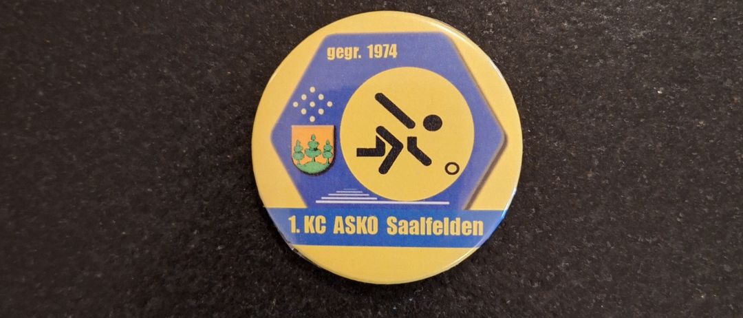 Eine gelbe kreisförmige Anstecknadel mit blauem Rand trägt die Aufschrift '1. KC ASKO Saalfelden' und ein Logo mit einem Skifahrer und dem Jahr 1974.