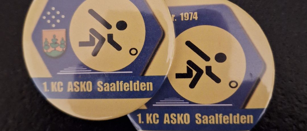 Zwei Abzeichen, gold und blau, aus dem Jahr 1974, mit einem Logo und dem Text '1. KC ASKO Saalfelden'. Eines hat 'gegr.' auf der linken Seite und das andere hat 'r.' auf der rechten Seite.