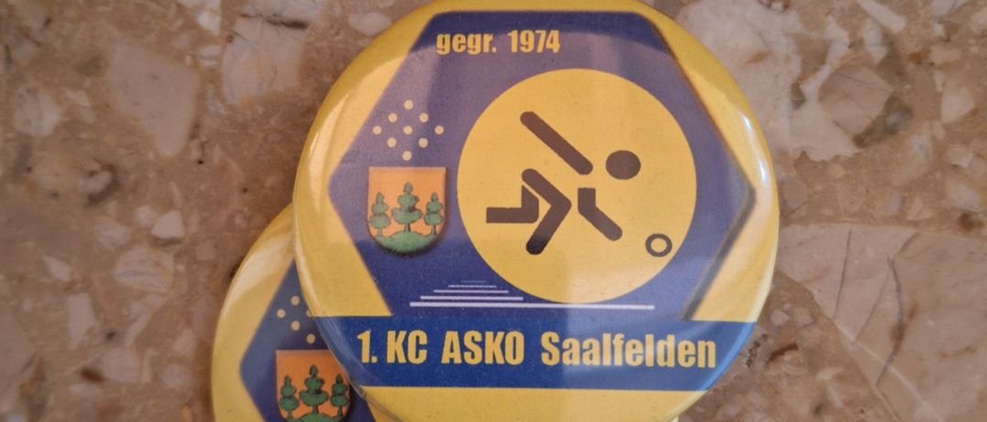 Zwei gestapelte gelbe Abzeichen mit '1. KC ASKO Saalfelden' und einer stilisierten Figur, dazu 'gegr. 1974'.